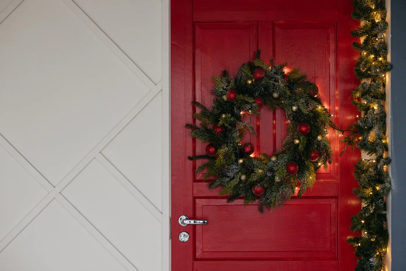 A beautifully decorated Christmas wreath hangs on a vibrant red door, lit by holiday lights.