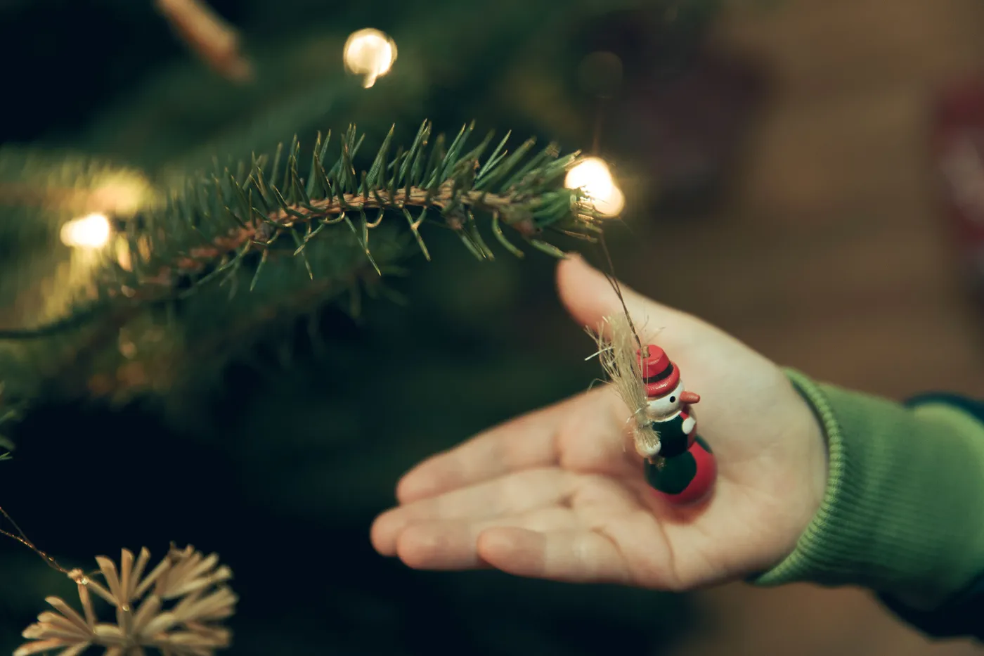 A close-up of a child placing a snowman ornament on a Christmas tree branch with lights.