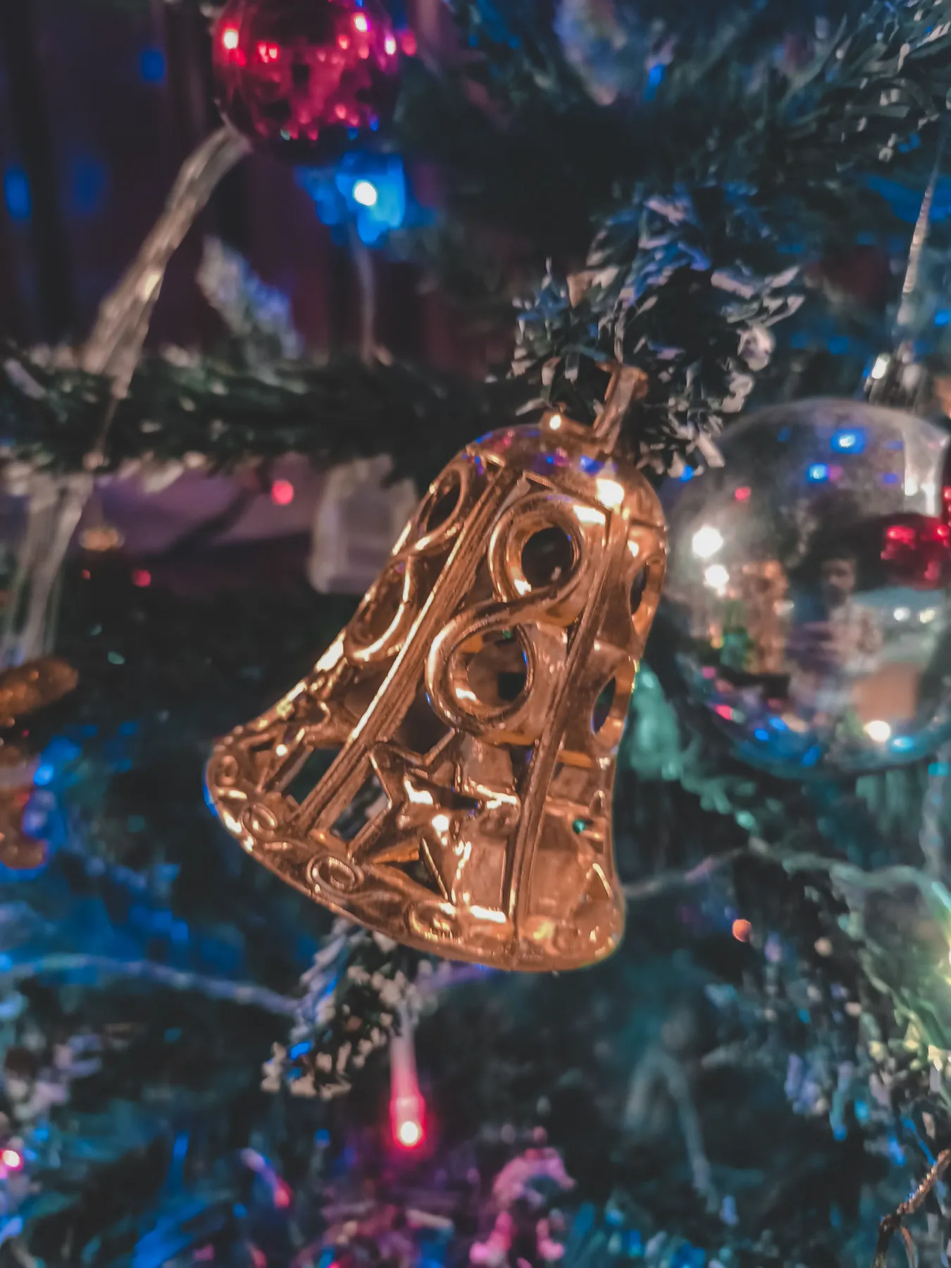 A close-up of a golden bell ornament hanging on a decorated Christmas tree with lights.