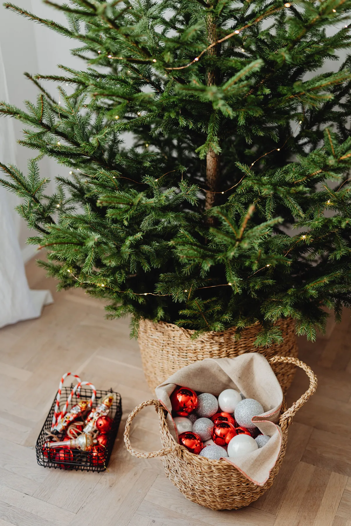 A decorated Christmas tree standing beside a basket filled with festive ornaments indoors.