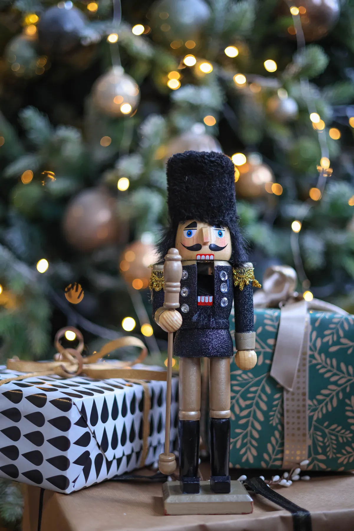 A decorative nutcracker soldier with holiday gifts against a Christmas tree background.