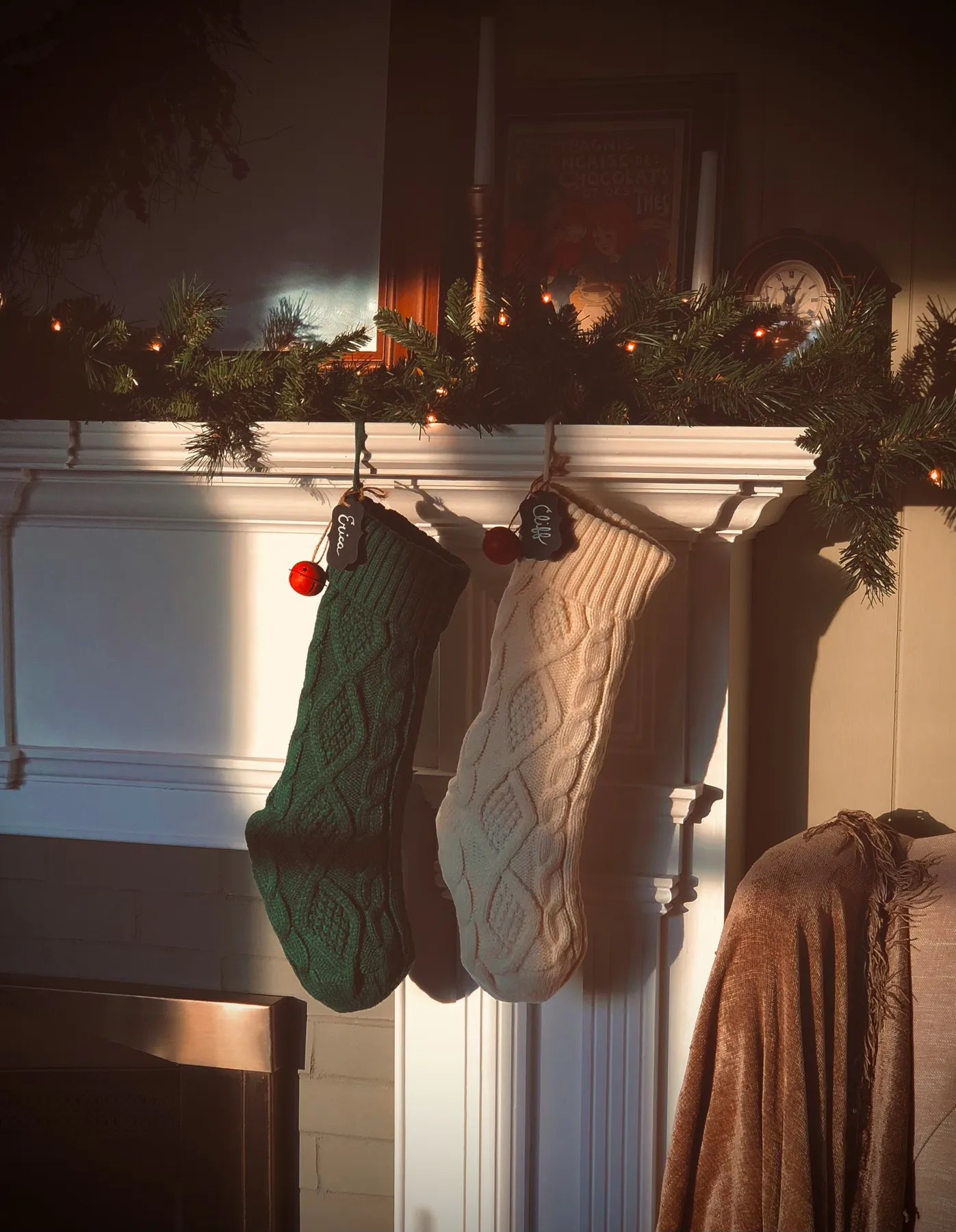 A green knitted holiday stocking and a white knitted holiday stocking sitting on an ornate white fireplace mantle as sun pours through a window and highlights the stockings. A red jingle bell and name