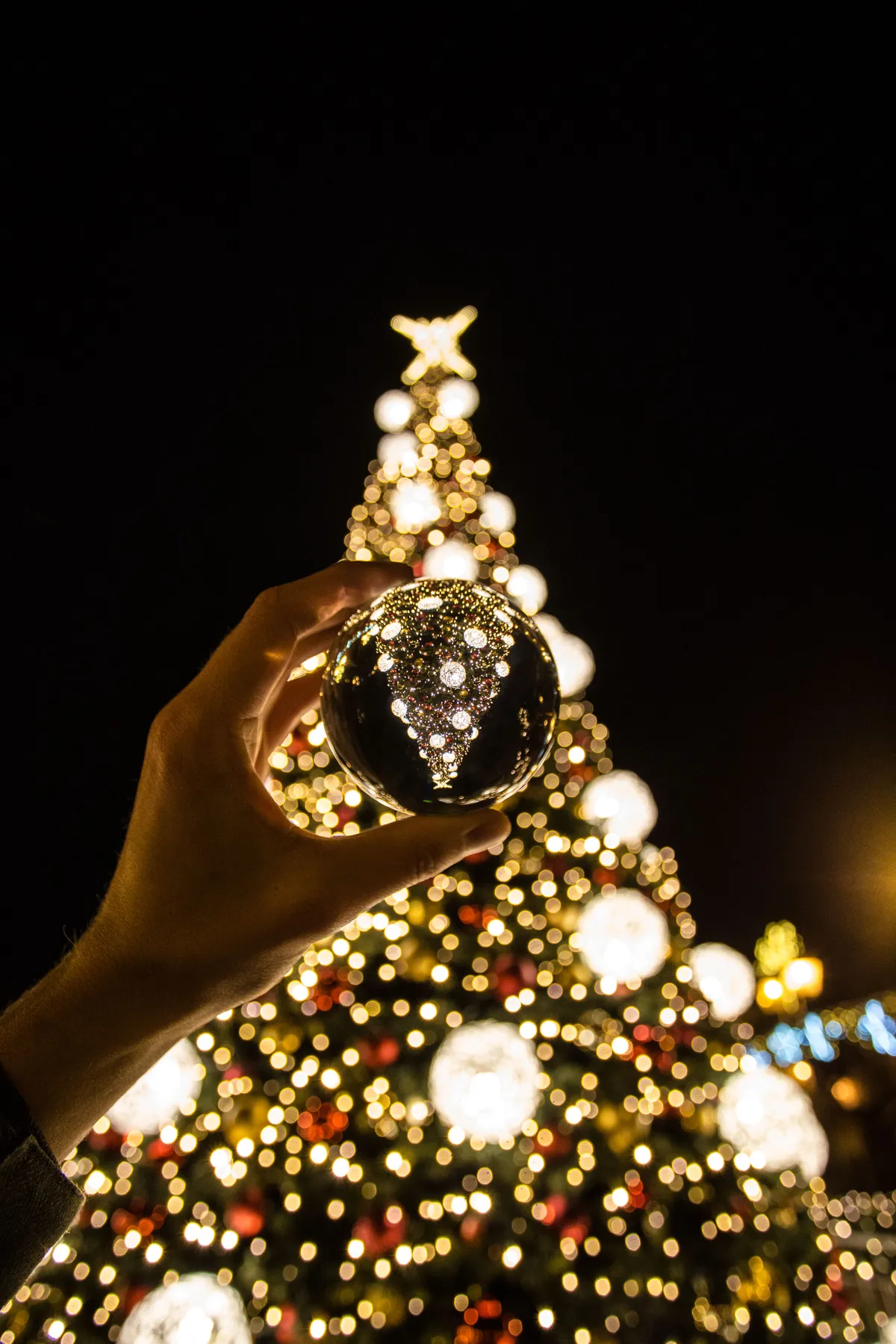 A hand holding a reflective ornament in front of a lit Christmas tree at night.