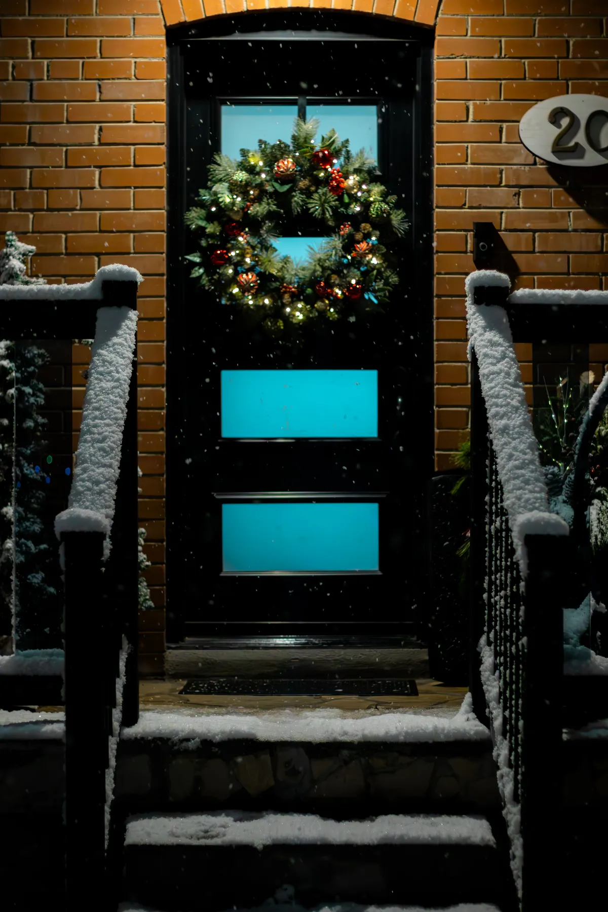 A warmly lit door in Toronto adorned with a festive Christmas wreath during snowfall.