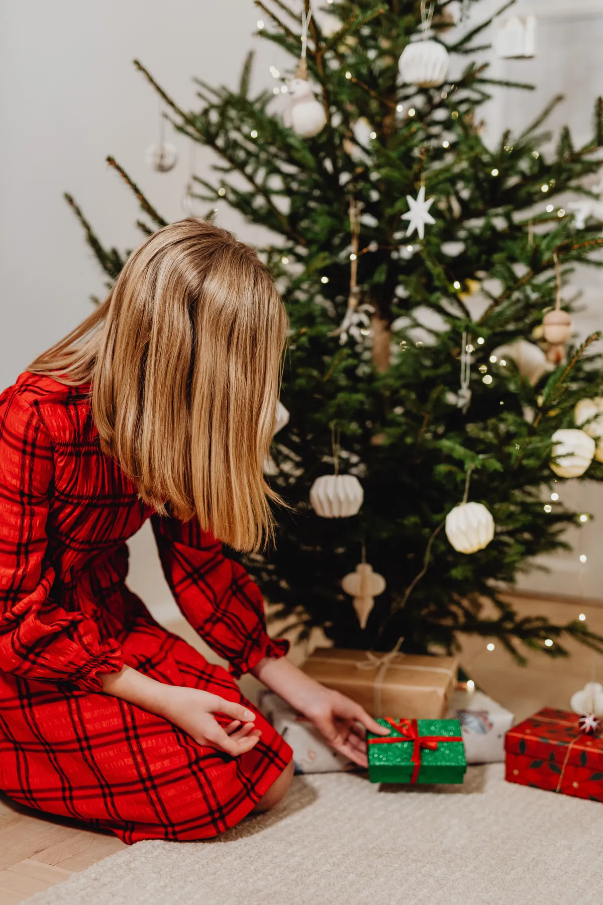 A woman in a red dress places presents under a decorated Christmas tree indoors.