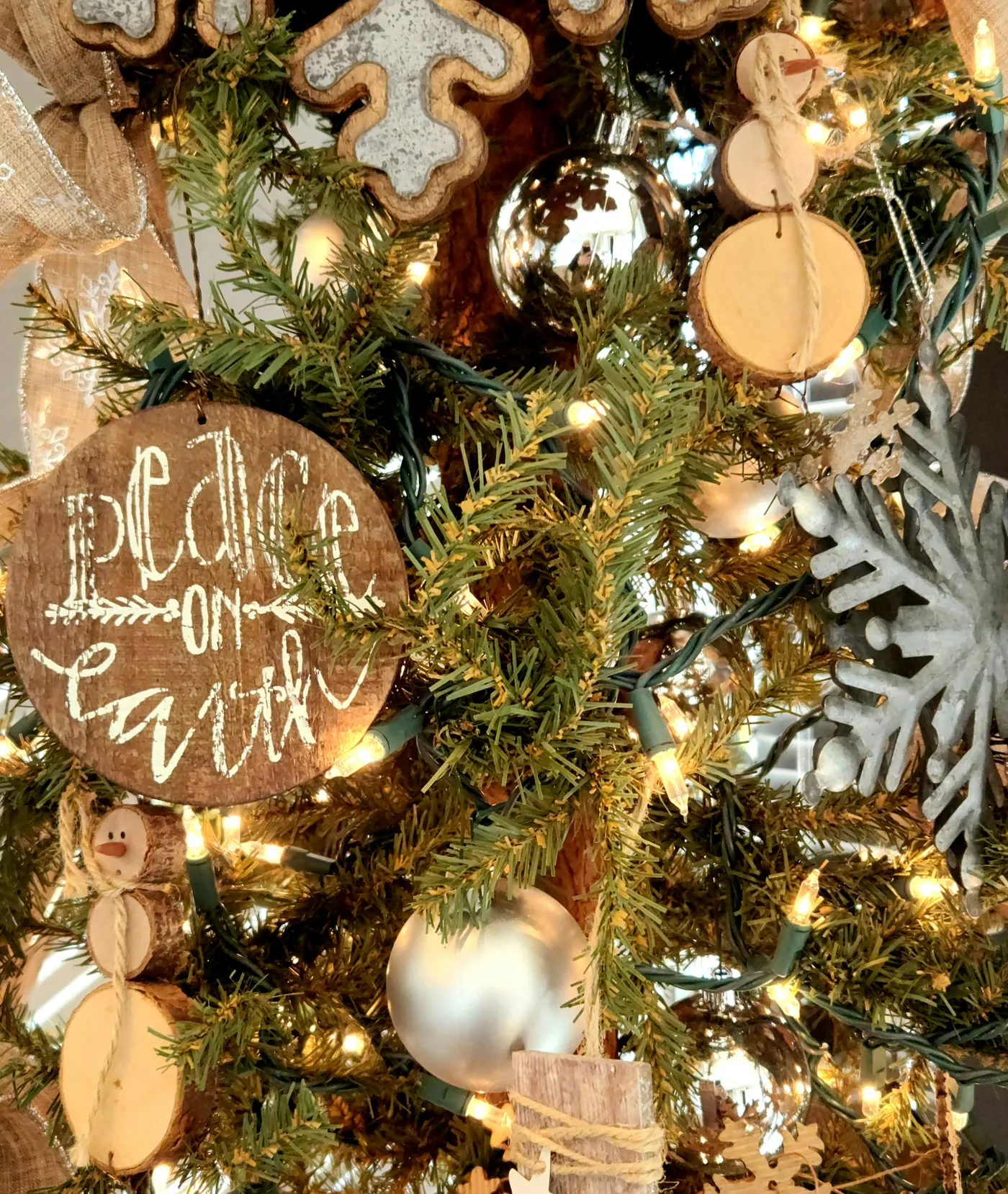 Christmas tree using wooden and tin ornaments. Focus is on a 'Peace on Earth' ornament and a snowflake metal ornament with white lights.