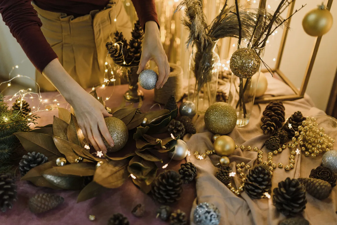 Hands arranging Christmas baubles and pine cones on a festive decorated table.
