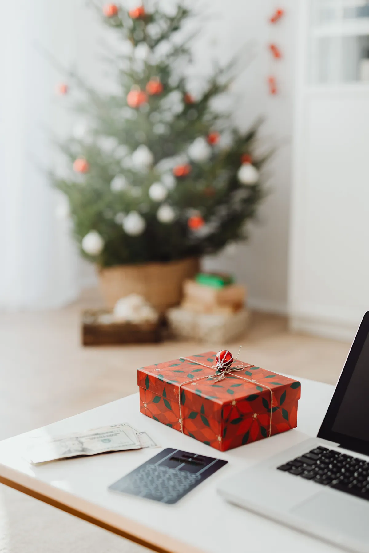 Red gift box on desk with laptop and Christmas tree in background, creating a cozy holiday atmosphere.