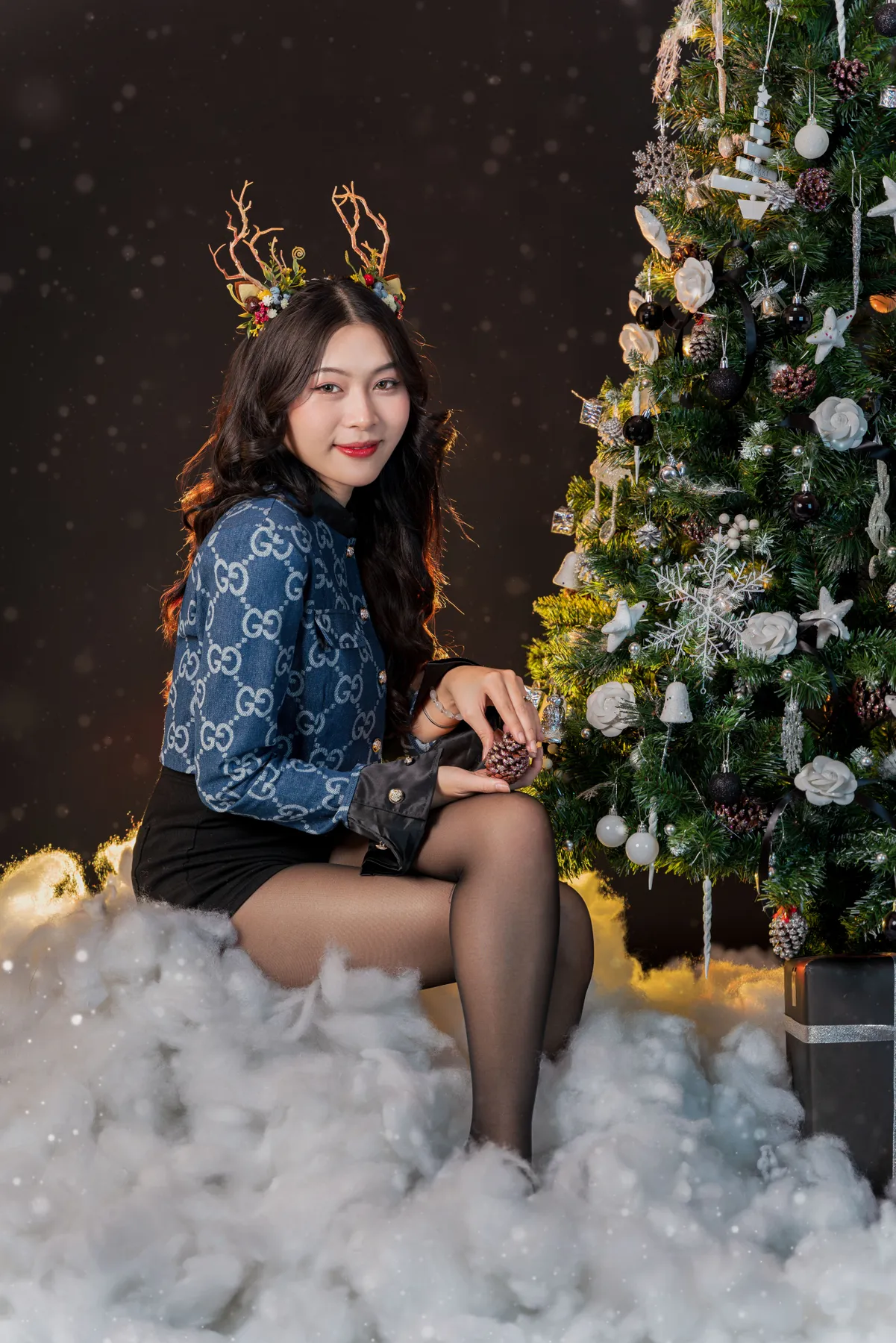 Woman wearing reindeer antlers sitting by a decorated Christmas tree with snow.